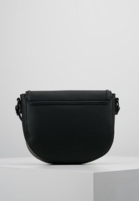 Borsa a tracolla in pelle nera con chiusura a pattina, fondo arrotondato e cinturini laterali regolabili, esposta su un piedistallo bianco.