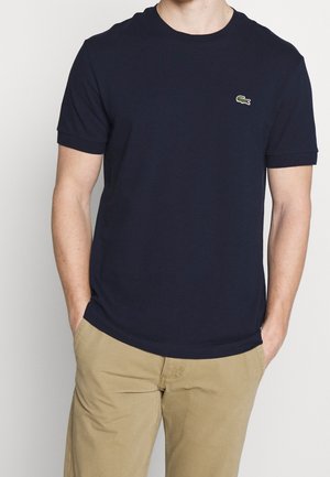 Mann trägt ein schlichtes dunkelblaues T-Shirt mit kleinem grünem Krokodil-Logo auf der Brust und beigen Hosen, Hände in den Taschen.