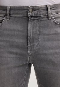 Jeans in denim grigio con una texture liscia, dotati di chiusura a bottoni, cinque tasche e dettagli di cucitura sottili sulle cuciture.