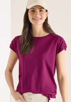 Donna sorridente che indossa un berretto beige, una maglietta a maniche corte magenta con spalle arricciate e pantaloni beige chiaro con dettagli a bottoni.