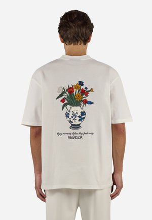 Άνδρας που φοράει λευκό μπλουζάκι με πολύχρωμη εικονογράφηση ανθοδέσμης και κείμενο "Απόλαυσε τις στιγμές πριν σβήσουν. PEGADOR" στην πλάτη.