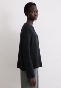 Czarny wełniany sweter z okrągłym dekoltem, opuszczonymi ramionami i luźnym krojem, noszony na szarych spodniach dresowych, na jasnym tle.