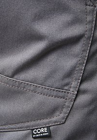 Pelēks audums ar gludu tekstūru, kuram ir kabata un šuvju detaļa. Apakšā ir birka ar uzrakstu "CORE by JACK & JONES".
