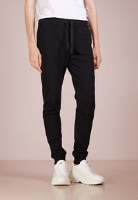 Pantalones jogger negros de tela suave con cinturilla ajustable, piernas ajustadas y paneles en contraste. Combinados con deportivas blancas gruesas.