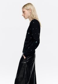 Melna adīta kardigāna, kas rotāta ar zaļa un sudraba akcentiem, pieguļoša piegriezuma, ar apaļu kaklu un garām piedurknēm, savienota ar melnu ādas svārku.