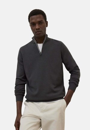 Hombre con un suéter gris oscuro con cremallera sobre una camisa blanca y pantalones beige claros, de pie con una mano en el bolsillo contra un fondo liso.