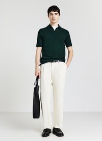 Giovane uomo che indossa un polo a maniche corte verde scuro, pantaloni chiari, cintura e scarpe nere, tiene una borsa tote nera, in piedi davanti a uno sfondo chiaro e semplice.