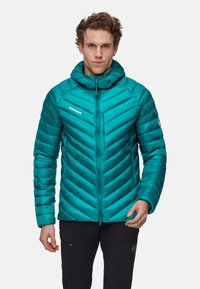 Mand iført en teal quiltet hættejakke og sorte bukser, der står imod en ensfarvet lys baggrund.