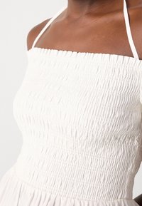 Tmavě pletěná osoba v bílých šatech s úzkými ramínky, zobrazená od krku po pas na jednobarevném pozadí.
