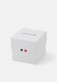 Caja rectangular blanca con tapa abatible, con el logo de "LACOSTE" y un cocodrilo verde. Rayas rojas y azules en la parte frontal inferior.