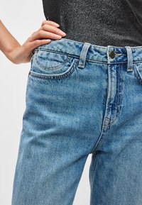 Ανοιχτό μπλε τζιν με άνετη εφαρμογή, που διαθέτει ραμμένες τσέπες, κλείσιμο με κουμπί και φερμουάρ, και ζώνη με θηλιές για ζώνη.