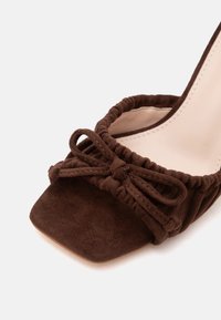 Chaussure ouverte en daim marron à bout carré avec bord froncé et nœud assorti sur la bride avant, montrant la doublure intérieure beige.