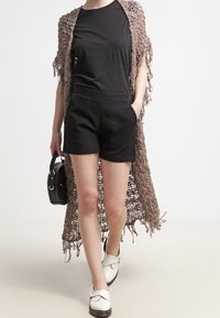 Svart kortärmad topp och högmidjade shorts, kombinerat med en lång, beige virk cardigan med fransdetaljer och vita skor.