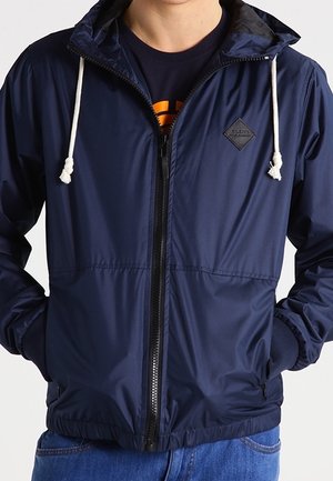 Mann trägt eine marineblaue Windjacke mit Reißverschluss, weißen Kordeln und elastischen Bündchen, Hände in den Taschen, über einem schwarzen und orangefarbenen Hemd.
