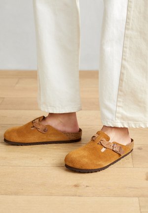 Braune Wildleder-Clogs mit runder Zehenpartie und Schnallenverschluss. Strukturiertes Obermaterial mit einem Korkfußbett, kombiniert mit crèmefarbenen, weit geschnittenen Hosen.