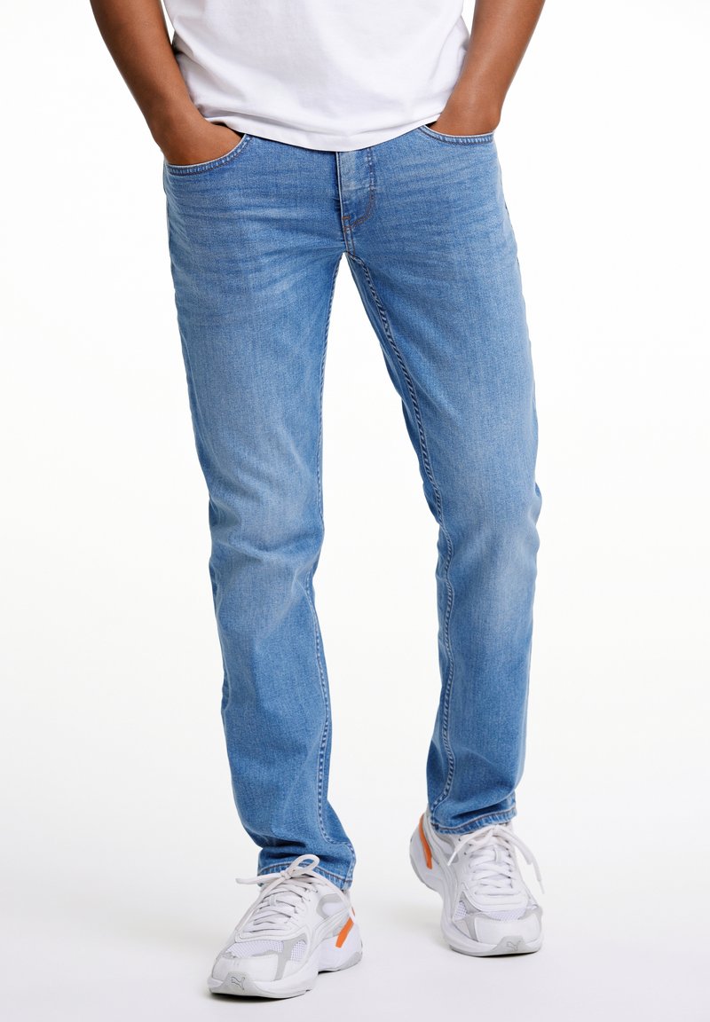 Light blue denim jeans with a straight-leg cut, featuring side pockets and subtle stitching, paired with white and gray athletic shoes.