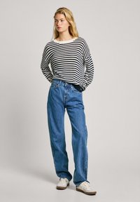 Pull en tricot noir et blanc à rayures, coupe oversize, associé à un jean bleu taille haute et des baskets blanches avec des accents marron clair.