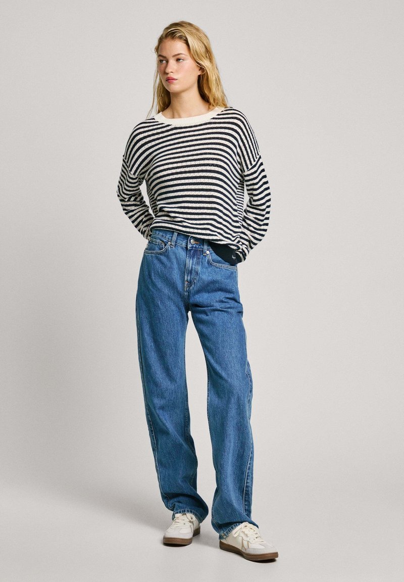 Pull en tricot noir et blanc à rayures, coupe oversize, associé à un jean bleu taille haute et des baskets blanches avec des accents marron clair.