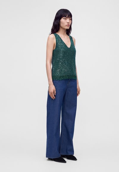 Femme debout portant un haut vert sans manches à sequins, un jean large bleu et des chaussures pointues noires sur un fond clair uni.