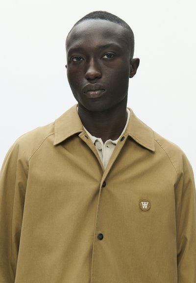 Jeune homme noir aux cheveux courts portant une veste marron clair boutonnée par-dessus une chemise blanche, faisant face à l'avant avec une expression neutre sur un fond uni.