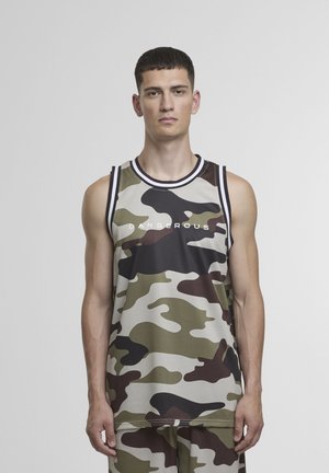 Mladý muž s krátkymi vlasmi, oblečený v maskáčovom tielku bez rukávov s nápisom „DANGEROUS“, stojaci pred jednoduchým svetlosivým pozadím.