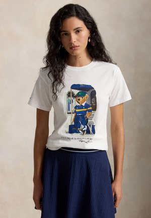 Femme aux longs cheveux bouclés porte un t-shirt blanc avec un ours en tenue polo bleue et jaune, assorti à une jupe texturée bleu marine.