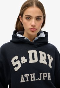 Navyblauwe hooded sweatshirt met een witte geborduurde logo "S&Dry ATH.JPN" op de voorkant, met een gestructureerde afwerking en een gelaagde lichtblauwe collared shirt daaronder.
