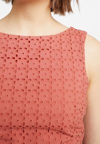 Haut sans manches couleur terre cuite avec un motif texturé de cercles et d'ouvertures linéaires, doté d'un col rond et d'une finition lisse.