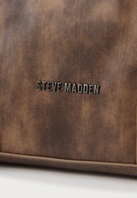 Brūna teksturēta ādas soma ar izcelta melna logo "STEVE MADDEN". Redzamie šuvumi gar apakšējo šuvi.