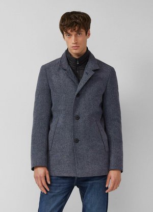 Young man wearing a gray wool blazer with black inner zip jacket and blue jeans, standing against a plain light gray background.