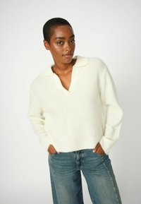 Weißer gestrickter Pullover mit einem tiefen V-Ausschnitt und einem Kragen, kombiniert mit blauen weiten Jeans. Texturiert, entspannte Passform.