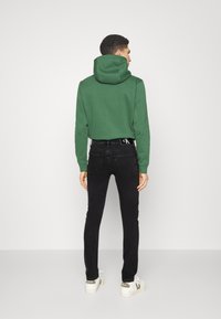 Sudadera verde con bolsillo frontal tipo canguro, combinada con jeans negros ajustados. Zapatillas blancas con detalle del logo. Diseño simple y casual.