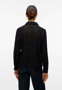 Blusa nera trasparente con righe verticali texturizzate e dettagli in perline sottili. Presenta un colletto e maniche lunghe, aderente in vita.