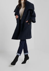 Granatowy płaszcz z mieszanki wełny z oversize'owym kołnierzem, dwurzędowym zapięciem i bocznymi kieszeniami, połączony z ciemnymi jeansami z podwiniętymi nogawkami i czarnymi botkami na obcasie.