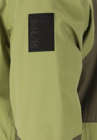 Grøn jakke med en tekstureret sort etiket med "SLOPE" og "STYLE" prægede. Stoffet ser glat og letvægtigt ud.