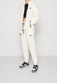 Bílý fleecový hoodie s černými akcenty v kombinaci s odpovídajícími bílými joggery s logem. Černobílé tenisky dokončují vzhled.