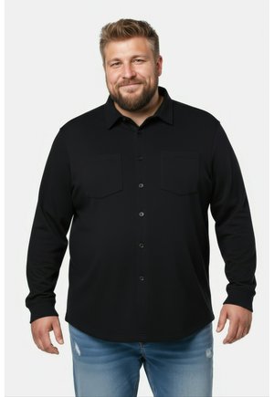 Lächelnder Mann mit kurzen Haaren und Bart, der ein schwarzes langärmliges Hemd mit Knöpfen und hellblaue Jeans trägt, steht vor weißem Hintergrund.