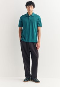 Žalsvai mėlynas polo marškinėlis su apykakle, trumpomis rankovėmis ir mažyčiu logotipu. Derinamas su tamsiomis kelnėmis ir juodais batais, kurie turi tvirtą padažą.