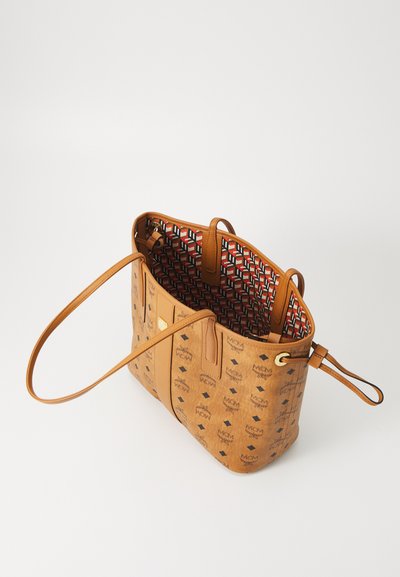 Cognackleurige MCM tote bag met zwarte logo patronen, rode geometrische stofvoering, twee schouderriemen en een goudkleurig metalen plaatje.