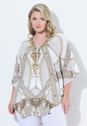 Blonde Frau trägt weiße Tunika mit goldenen und schwarzen, kunstvollen barocken Mustern, kombiniert mit weißen Hosen, steht vor einer weißen Wand.