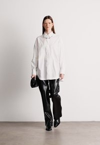 Λευκό κορυφαίο πουκάμισο με oversized γραμμή και υφή μπροστά, συνδυασμένο με λαμπερό μαύρο παντελόνι με σχέδιο κροκό, και μία μικρή μαύρη τσάντα.