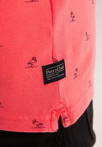 Korallrosa skjorta med små svarta flamingomönster, texturerad tyg och ett synligt märkesetikett med "Petrol Ind" nära botten.