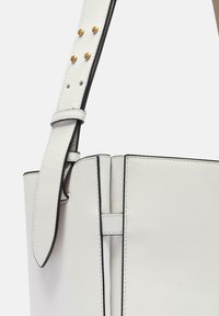 Biała skórzana torba typu bucket o sztywnym kształcie, podkreślona czarnymi krawędziami i złotymi ćwiekami na regulowanym pasku.