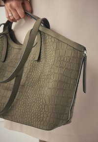 Sac à main vert olive avec texture de crocodile embossée, doté de longues poignées, d'une fermeture éclair et d'accents gris clair le long des bords.