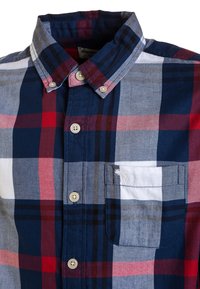 Koszula zapinana na guziki w granatowo-czerwono-szary wzór w kratkę. Posiada kołnierzyk, zapięcie na guziki z przodu oraz kieszeń na piersi z białym detalem.