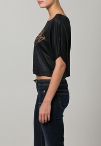 Černý cropped top z měkké, hladké látky. Na přední straně má zlatý geometrický vzor. Doplněno tmavě modrými džíny.