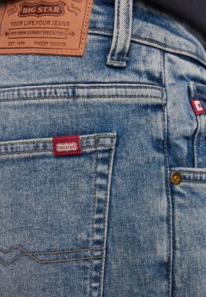 Niebieskie dżinsy z denimu z skórzaną łatą marki, czerwoną metką i mosiężnym sprzętem, widocznymi szwami i wyblakłą teksturą.