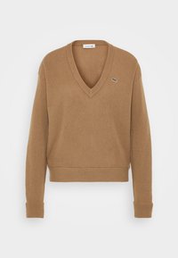 Brun V-ringad tröja i stickad textur, med långa ärmar och ribbad nederkant. Litet grönt logotypaccent på vänster bröst.