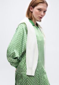 Jeune femme aux cheveux rouges courts portant une robe verte à motif et un pull blanc posé sur ses épaules, sur un fond uni.