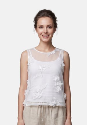 Giovane donna sorridente, indossa un top bianco senza maniche e trasparente con applicazioni floreali 3D e pantaloni beige su uno sfondo chiaro uniforme.